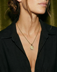 Close-up of a person wearing a black shirt with a gold necklace featuring a pendant against a green curtain background.
