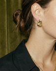 Close-up of a person wearing gold hoop earrings with a blurred green curtain background