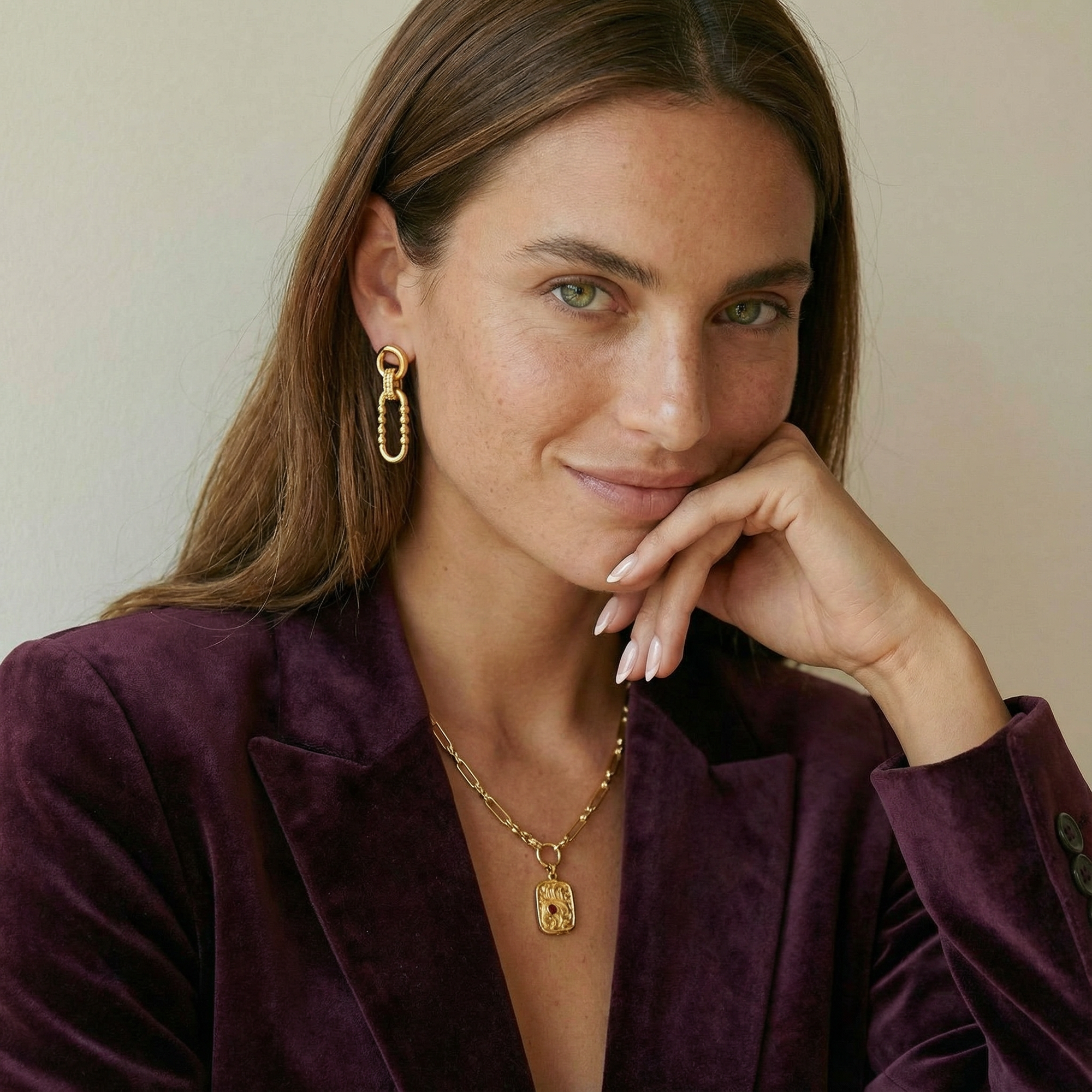 Woman wearing gold earrings and necklace, posing with hand on chin against a neutral background