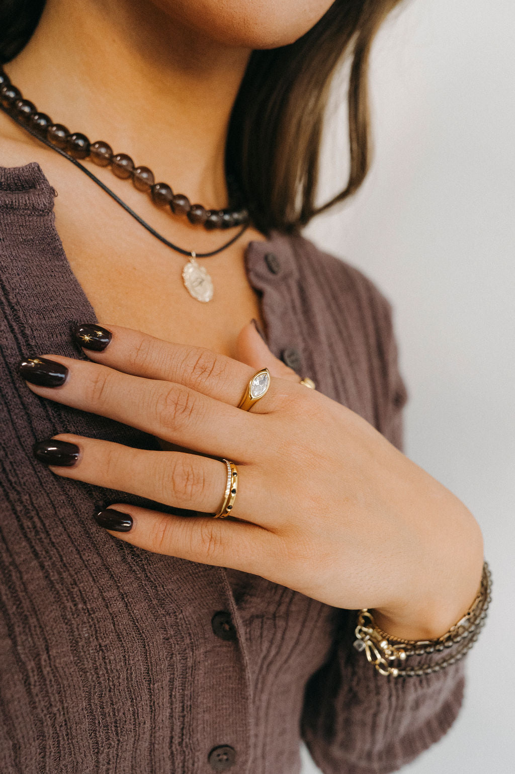 Close-up of a person wearing multiple necklaces, rings, and a bracelet on a neutral background