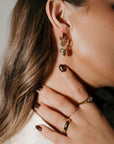 Close-up of a person wearing gold hoop earrings and rings with a neutral background