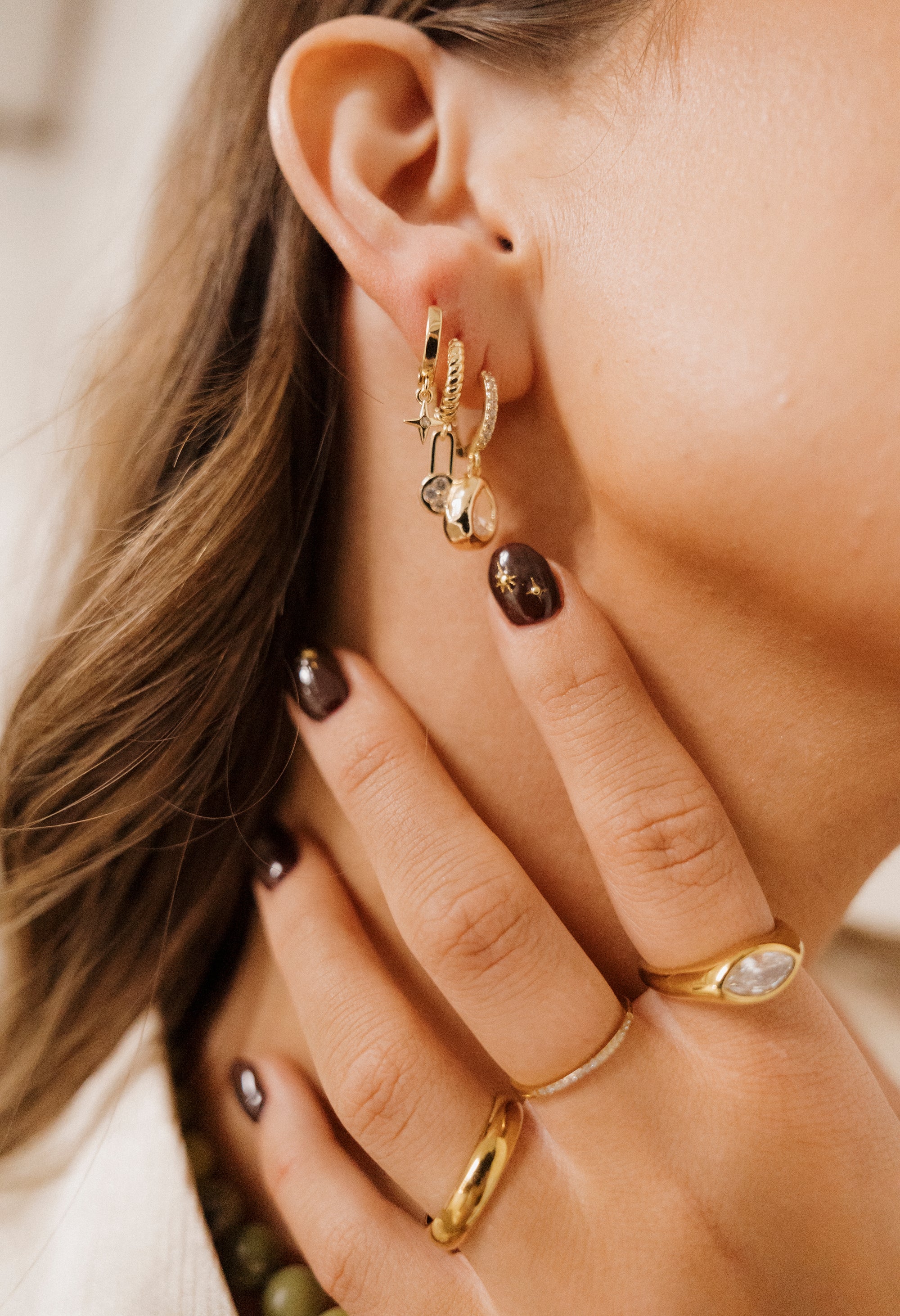 Close-up of a person's ear with gold hoop earrings and rings on a neutral background