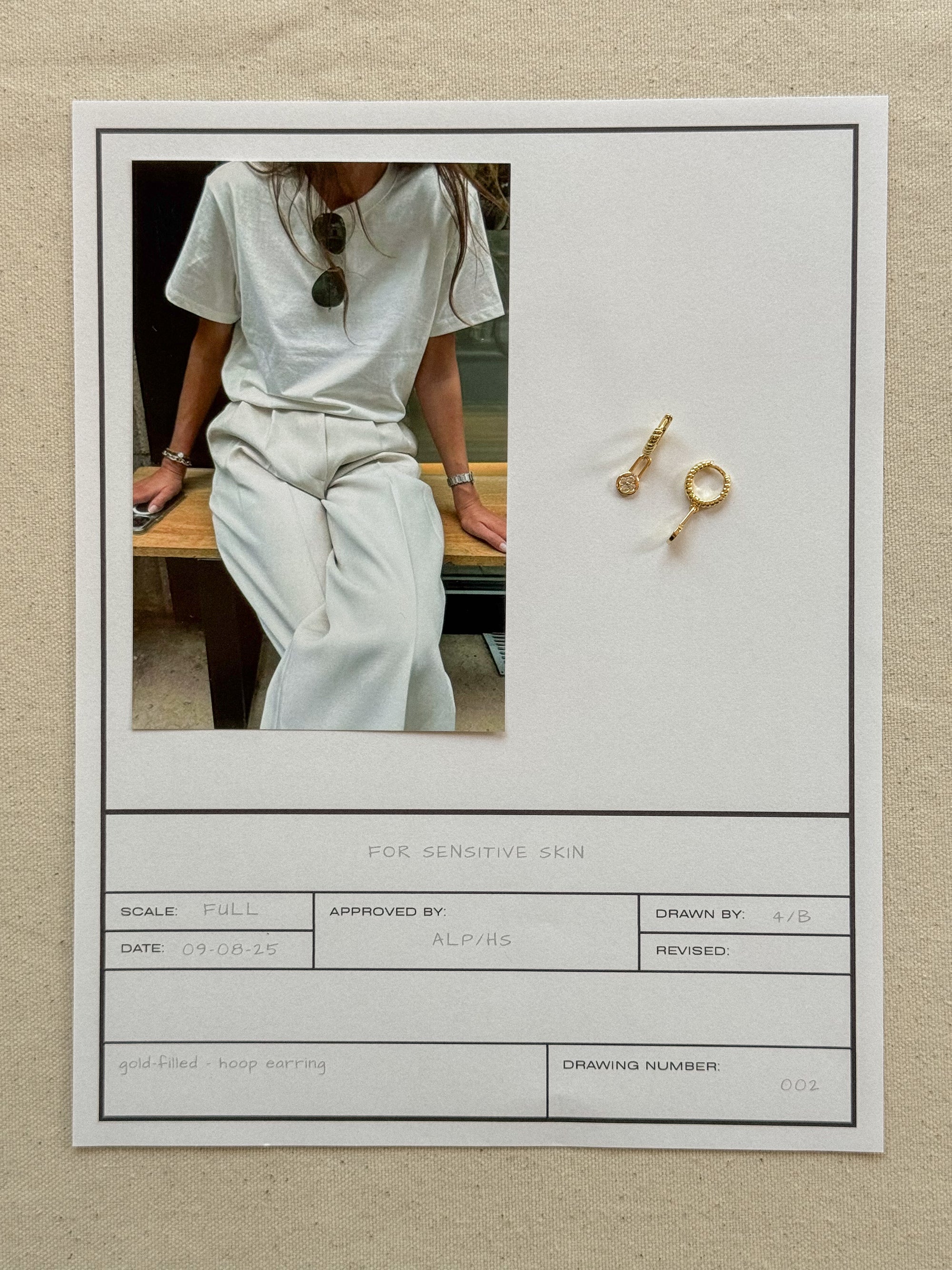 Person wearing a white outfit with sunglasses, sitting on a bench, and gold earrings displayed on a neutral background.