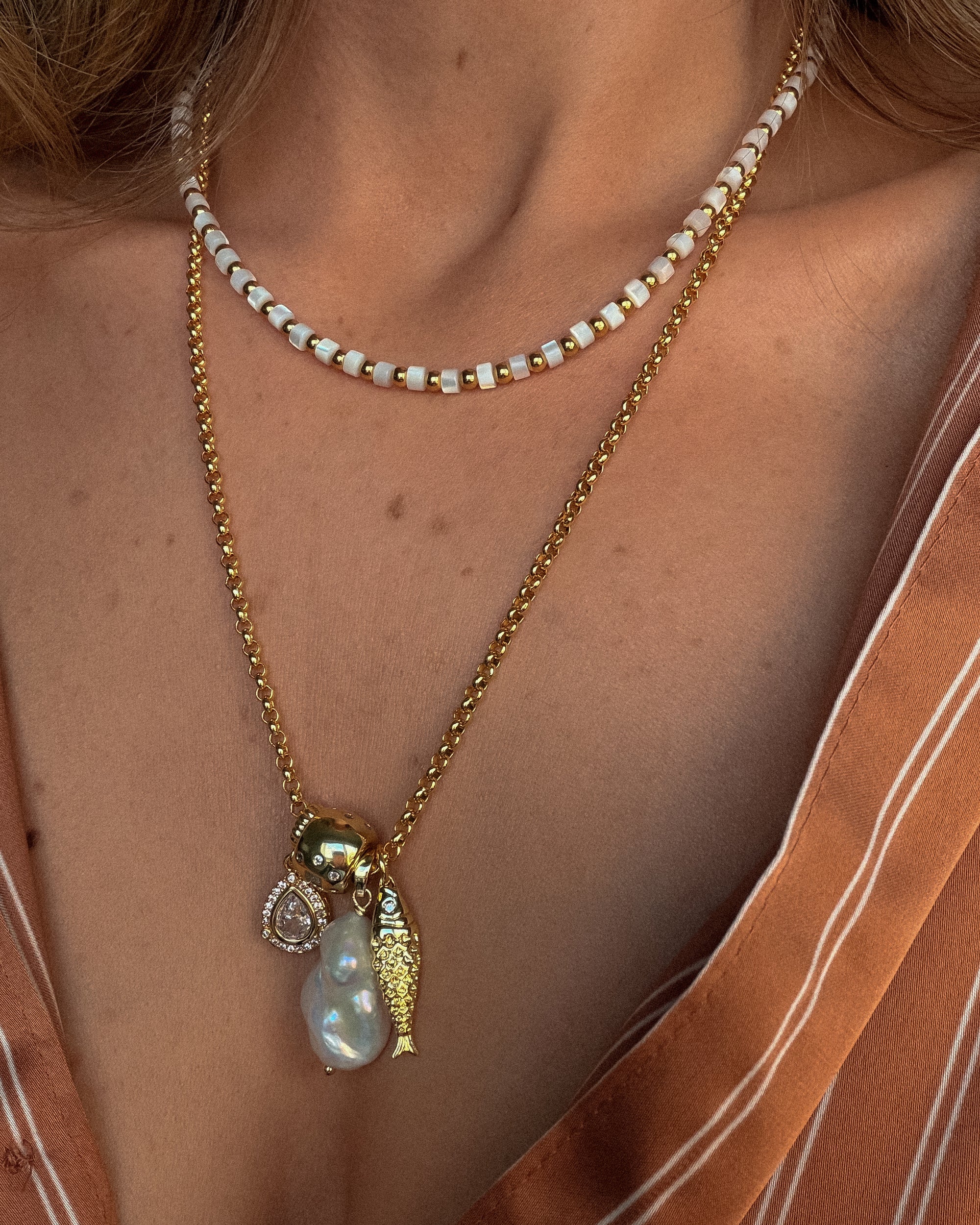 Close-up of a person wearing a gold necklace with a pearl pendant and fish charm.