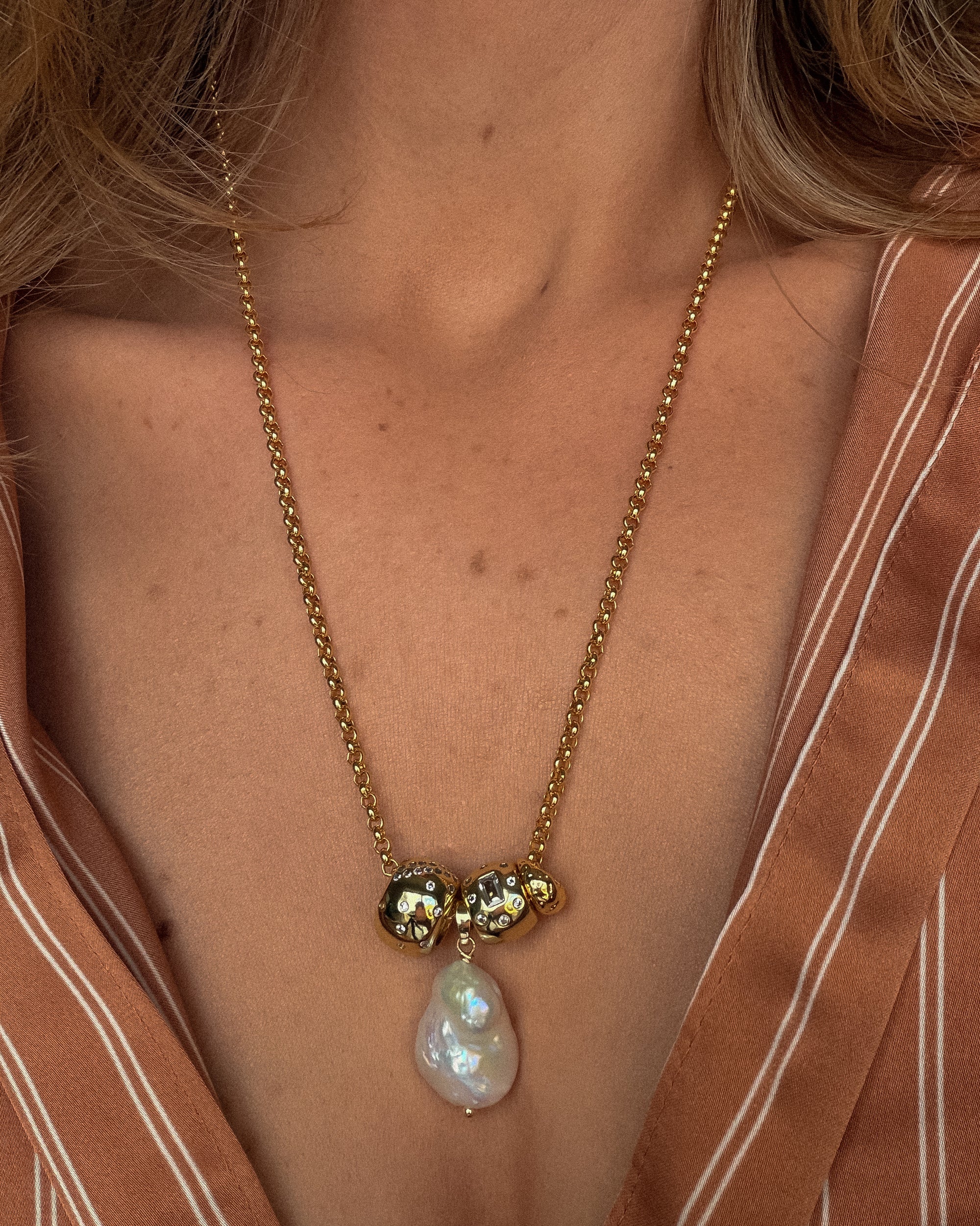 Gold necklace with a pearl pendant worn by a person in a brown striped garment.