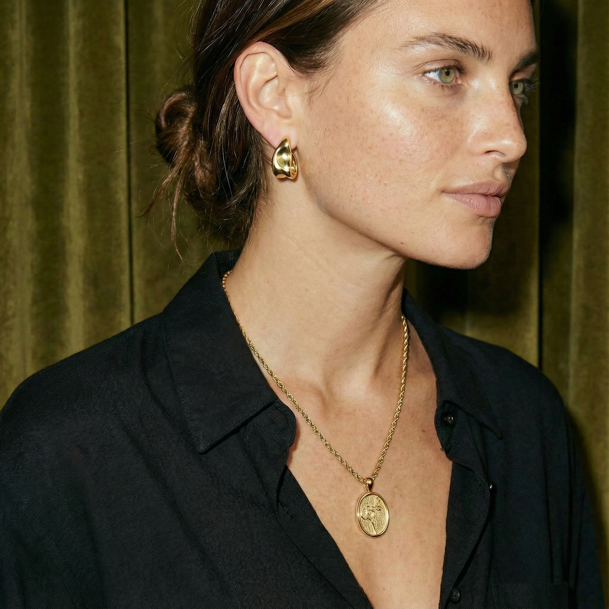 Woman wearing gold earrings and necklace against a green curtain background
