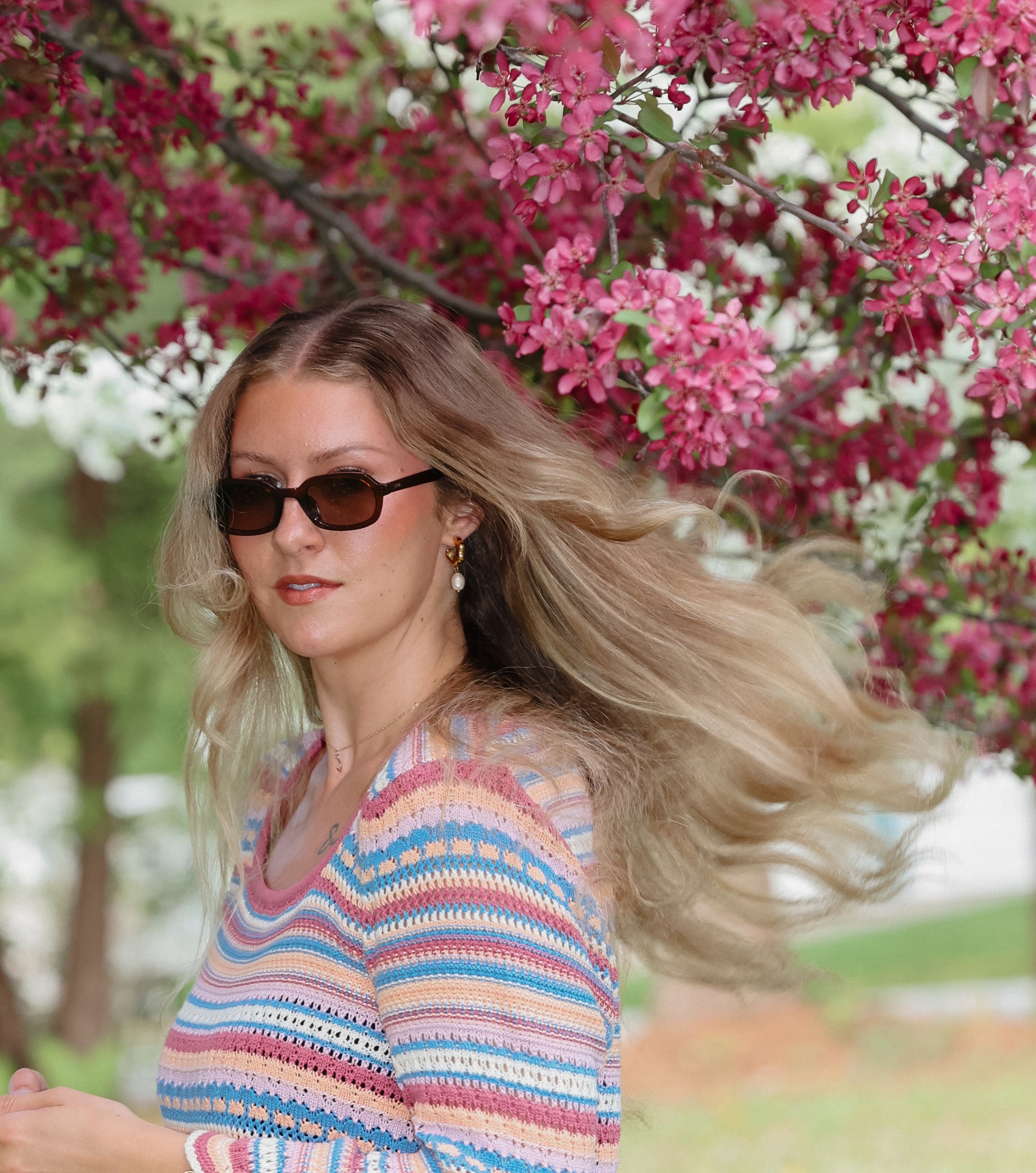 Woman with sunglasses and a colorful striped sweater standing in front of pink flowering trees.