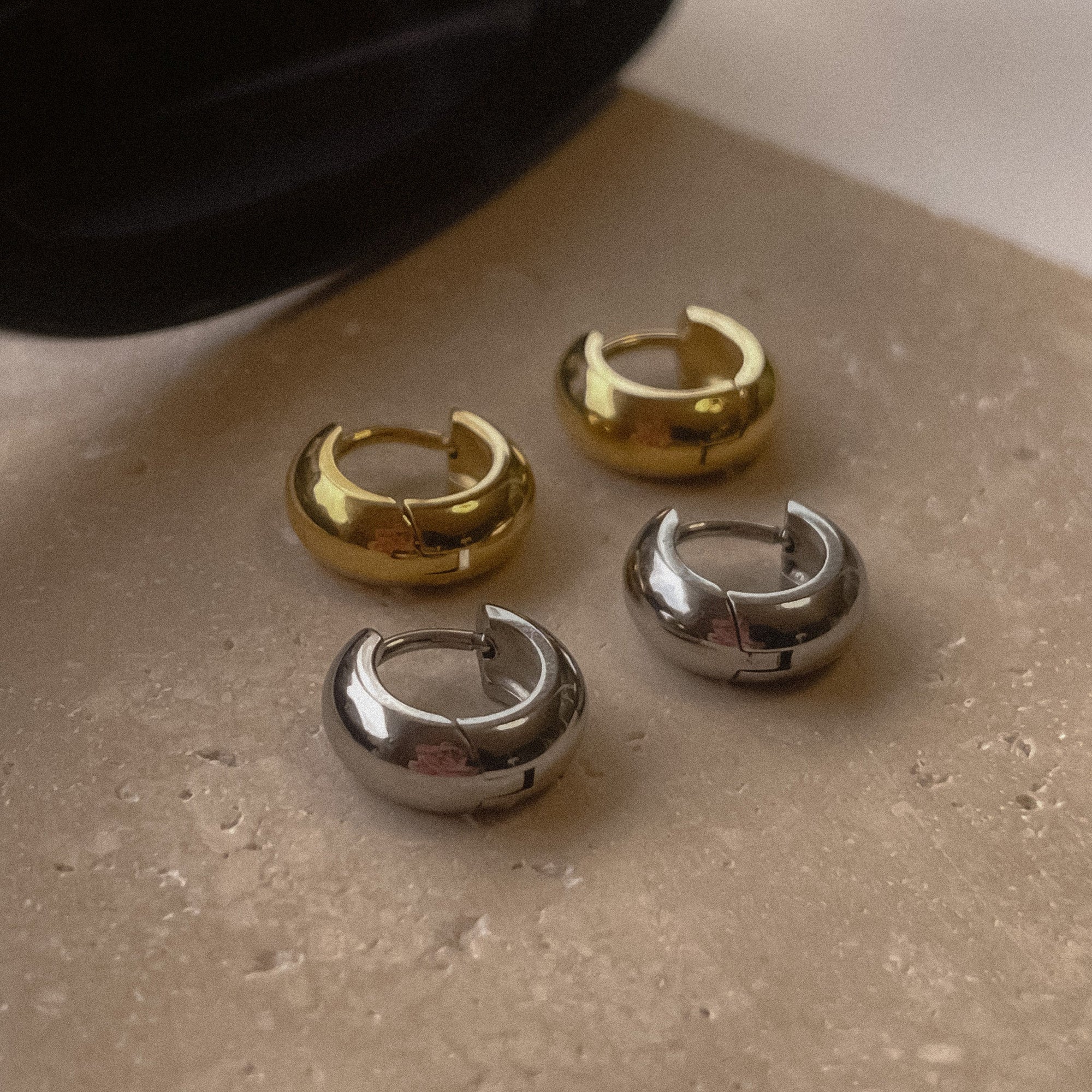 A pair of shiny gold hoop earrings and a pair of shiny silver hoop earrings on a stone with sunglasses in the background.