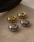 A pair of shiny gold hoop earrings and a pair of shiny silver hoop earrings on a stone with sunglasses in the background.
