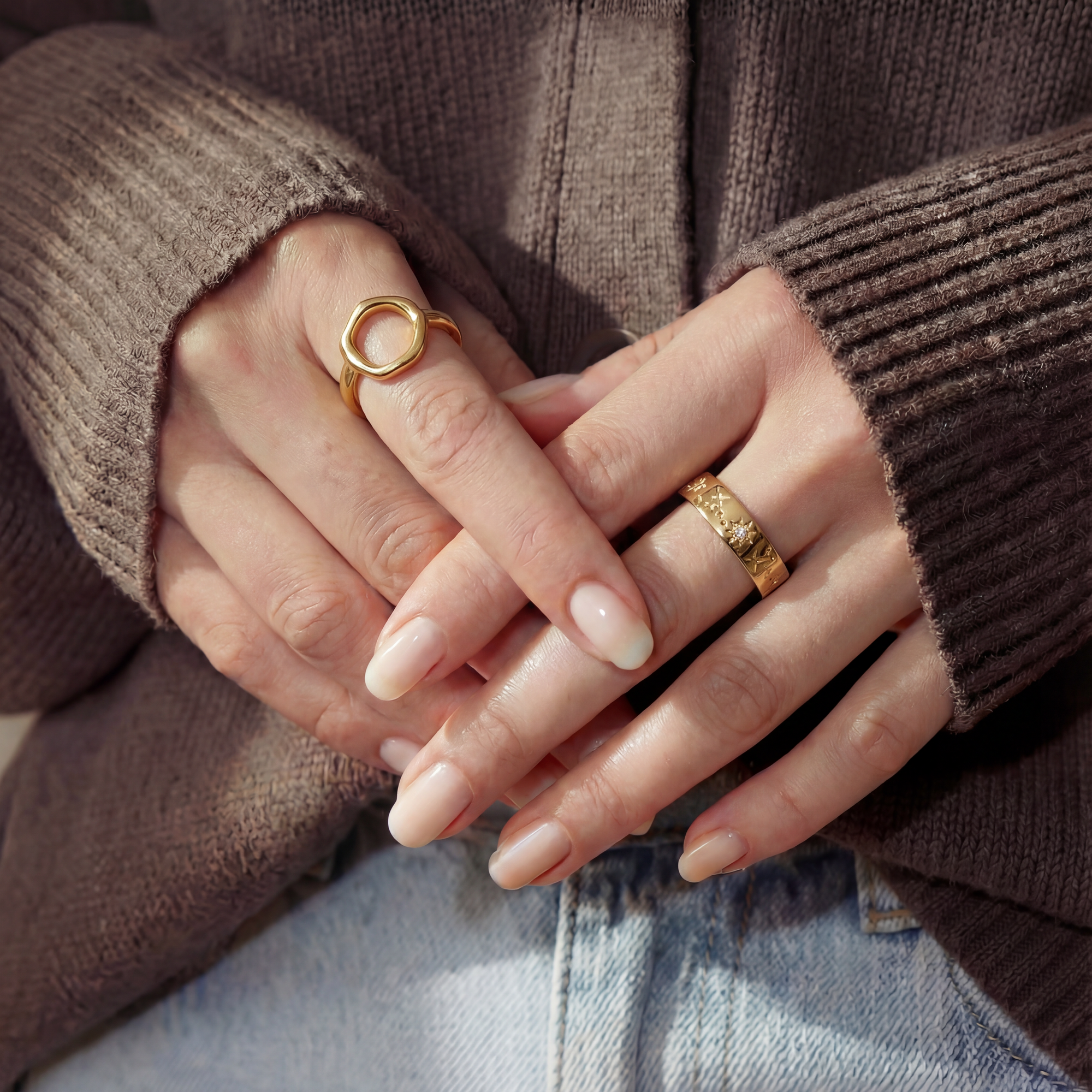 Close-up of hands with gold rings wearing a brown sweater and blue jeans.
