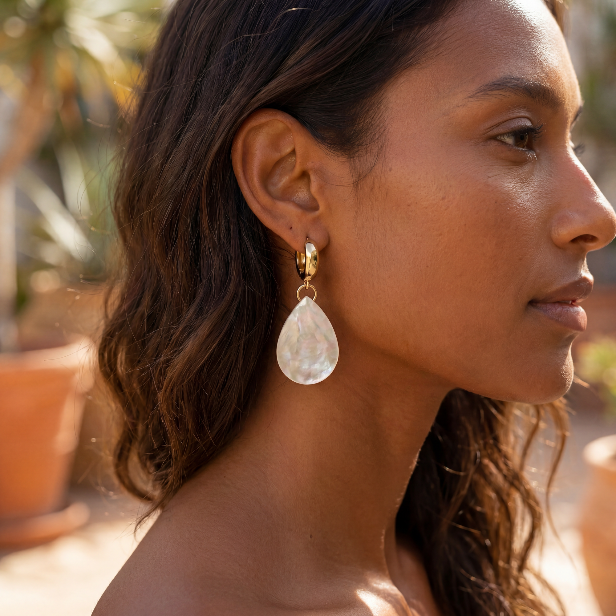 dark complexion woman with beach brown wavy hair wearing large shell tear drop gold plated hoops. Beach background.