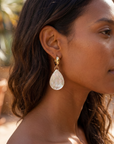 dark complexion woman with beach brown wavy hair wearing large shell tear drop gold plated hoops. Beach background.